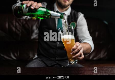 Un homme verse de la bière de blé légère dans une bouteille en verre, en bulles et en mousse. Homme dans un gilet et une chemise Banque D'Images