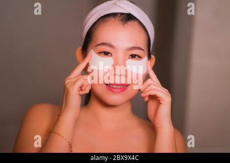 Jeune femme chinoise asiatique belle et heureuse souriant au miroir de salle de bains appliquant des timbres hydratants sous les yeux pour réparer les cercles sombres dans le beaut Banque D'Images