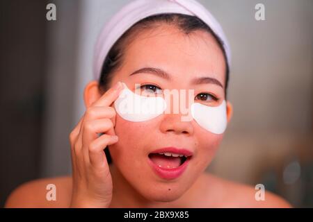 Jeune femme chinoise asiatique belle et heureuse souriant au miroir de salle de bains appliquant des timbres hydratants sous les yeux pour réparer les cercles sombres dans le beaut Banque D'Images