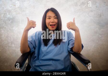 Portrait isolé de jeune femme asiatique chinoise belle et heureuse à l'hôpital robe de patient assis sur fauteuil roulant sourire positif de récupération de ACC Banque D'Images