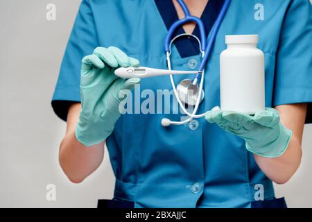 femme médecin avec stéthoscope portant un masque de protection et des gants en latex sur fond gris clair tenant thermomètre et peut pot de pilules de médicaments Banque D'Images