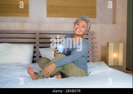 Portrait à la maison de femme asiatique mature et attrayante avec des cheveux gris assis sur le lit boire un café détendu sourire heureux comme milieu a Banque D'Images