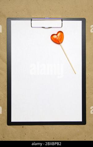 La tablette avec un clip pour le clip de papier avec une feuille blanche a4 repose sur l'arrière-plan du papier brun froissé. Sur le dessus se trouve un lollipop de caramel de rouge orange en forme de coeur. Place pour le texte et la mise en page pour la conception. Banque D'Images