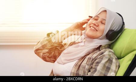 Jeune femme musulmane asiatique heureuse portant le hijab écoutant de la musique à partir d'un casque tout en étant assise sur une chaise confortable, les yeux fermés appréciant son temps Banque D'Images