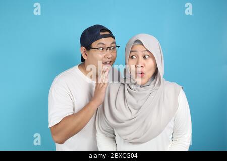 Homme couvrant sa bouche et chuchotant secret à son oreille de femme, femme musulmane asiatique a l'air choqué et excité, ouvrant les yeux et la bouche largement avec surpr Banque D'Images