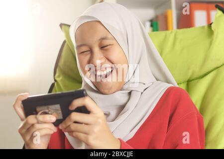 Portrait de jolie fille asiatique musulmane adolescente portant hijab jouant à des jeux en ligne sur le téléphone, jeune fille heureuse et excitée faisant des activités de loisirs Banque D'Images