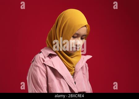 Portrait d'une jeune fille musulmane asiatique adolescente triste, portant le hijab, regardant vers le bas et déprimée, debout sur fond rouge Banque D'Images