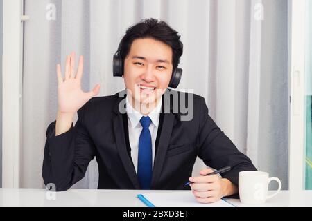 Travailler à domicile, un jeune homme d'affaires asiatique porte son costume pour la vidéoconférence ou facetime il souriant assis sur le bureau avec un casque et lève la main Banque D'Images