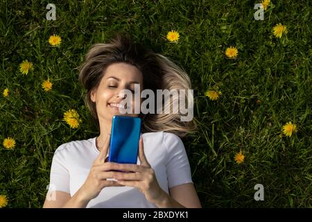 vue de dessus d'une jeune femme blonde, couchée sur de l'herbe verte et utilisant un smartphone Banque D'Images