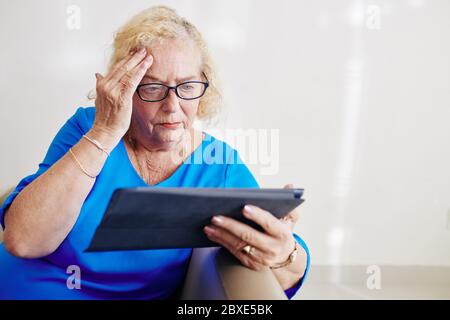 Femme âgée confuse dans des lunettes lisant des nouvelles troublantes sur un ordinateur tablette Banque D'Images