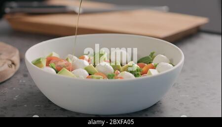 l'homme ajoute l'huile dans la salade avec le chou vert, l'avocat, la mozzarella et les tomates dans un bol blanc sur le comptoir de la cuisine Banque D'Images