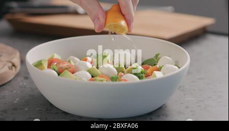 l'homme presse le citron orange dans la salade avec du chou vert, de l'avocat, de la mozzarella et des tomates dans un bol blanc sur le comptoir de la cuisine Banque D'Images