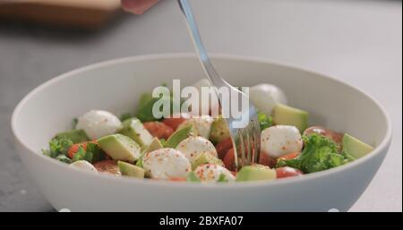 l'homme a le goût de la salade avec du kale, de l'avocat, de la mozzarella et des tomates dans un bol blanc sur le comptoir de la cuisine Banque D'Images