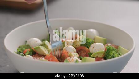 l'homme a le goût de la salade avec du kale, de l'avocat, de la mozzarella et des tomates dans un bol blanc sur le comptoir de la cuisine Banque D'Images