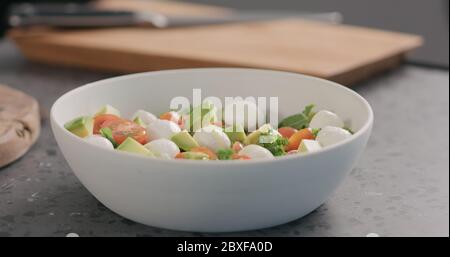 l'homme ajoute l'huile dans la salade avec le chou vert, l'avocat, la mozzarella et les tomates dans un bol blanc sur le comptoir de la cuisine Banque D'Images