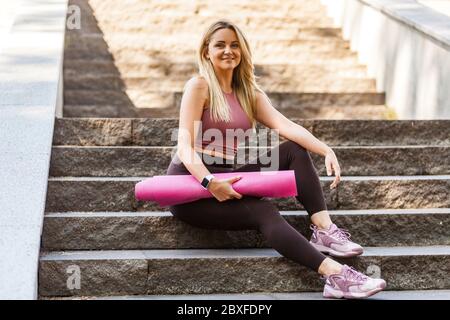 Bonne tenue blonde fille dans le pantalon de yoga assis sur les escaliers, tenant le tapis et souriant avec heureux expression satisfait, se détendre prendre une pause après le sport tra Banque D'Images