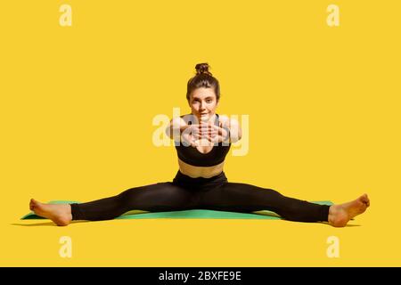 Mettre la femme de gymnastique avec un pain de cheveux dans les vêtements de sport serrés assis sur le tapis avec les jambes et les mains étirant, en se réchauffant, faisant des exercices de flexibilité sportive Banque D'Images