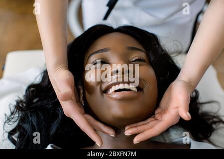 Gros plan des mains de la femme cosmetologiste faisant un massage spécial du visage à une jeune femme africaine souriante allongé sur un canapé dans un salon de beauté. Concept de Banque D'Images