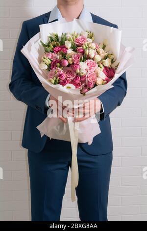 un jeune homme blond en costume bleu tient en main un bouquet géant de roses blanches-roses et d'eustoma sur le fond clair Banque D'Images