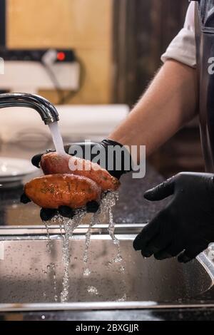 Gros plan d'un homme méconnaissable en gants de latex lavant des patates douces sous l'eau du robinet Banque D'Images