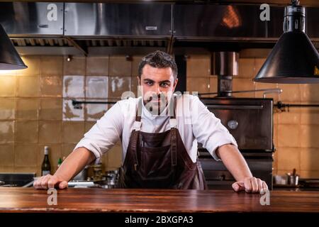 Portrait d'un chef à la barbe confiant dans un tablier en cuir debout sur un comptoir en bois dans une cuisine ouverte Banque D'Images