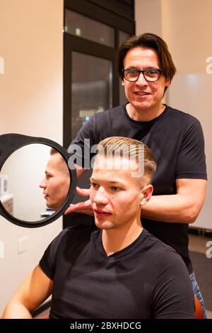 Coiffeur montre jeune homme après avoir coupé ses cheveux avec un miroir sa nouvelle coiffure dans son salon de coiffure Banque D'Images