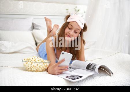 Une jeune fille se couche au lit après avoir dormi et navigue sur les réseaux sociaux au téléphone. Temps libre en dehors de l'école. Vie en quarantaine. Banque D'Images