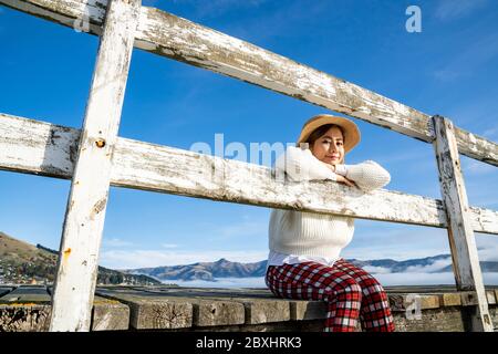 Une femme asiatique magnifique se reposant est regarde la baie de mer avec des maisons et des bateaux lors d'une journée ensoleillée à Childrens Bay, Akaroa, Canterbury, Nouvelle-Zélande. Détente Banque D'Images