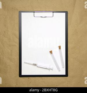La tablette avec un clip pour le clip de papier avec une feuille blanche a4 repose sur l'arrière-plan du papier brun froissé. Sur le dessus se trouve une seringue médicale et deux tubes vides transparents avec bouchons. Place pour le texte et la mise en page pour la conception. Banque D'Images