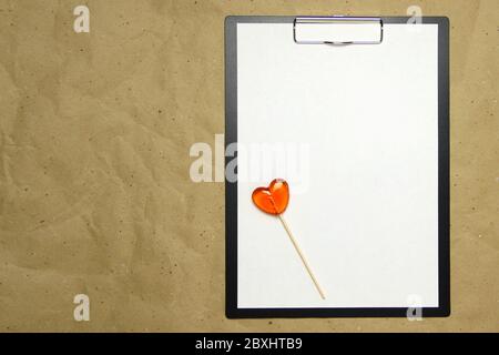 La tablette avec un clip pour le clip de papier avec une feuille blanche a4 repose sur l'arrière-plan du papier brun froissé. Sur le dessus se trouve un lollipop de caramel de rouge orange en forme de coeur. Place pour le texte et la mise en page pour la conception. Banque D'Images