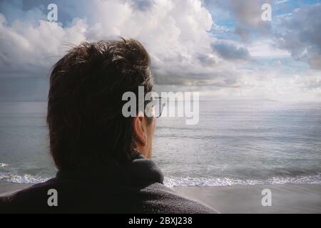 Arrière homme tête. Homme aux cheveux foncés avec lunettes de soleil noires et pull à col roulé regarde à l'horizon depuis une plage de l'Atlantique. Gros plan portrait en extérieur avec Banque D'Images