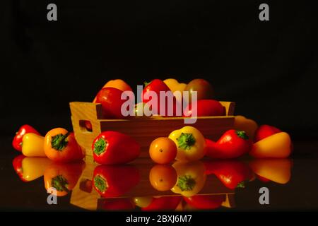 Quelques petits poivrons de couleurs rouge et jaune dans une petite boîte en bois qui est sur une table où les poivrons sont réfléchis. Fond sombre, nourriture sombre Banque D'Images