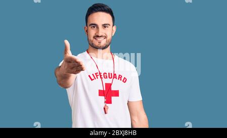 Jeune homme beau portant un maître-nageur uniforme souriant amical offrant poignée de main comme accueil et accueil. Entreprise réussie. Banque D'Images