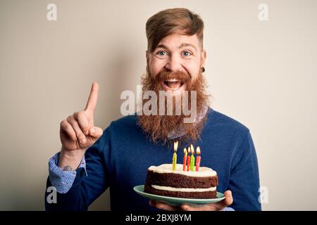 Homme irlandais à tête rouge avec barbe tenant gâteau d'anniversaire avec bougies allumées sur fond isolé surpris par une idée ou une question pointant du doigt wi Banque D'Images