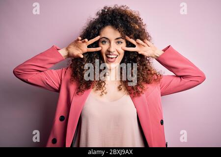 Jeune femme d'affaires avec des cheveux bouclés et piercing portant une élégante veste Faisant le symbole de la paix avec les doigts sur le visage, souriant et joyeux affichage Banque D'Images