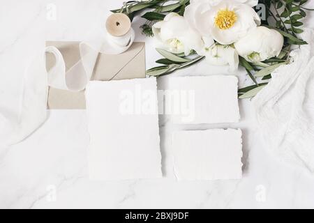 Papeterie de mariage, scène de vie. Enveloppe, cartes de vœux, maquettes en papier de coton. Bouquet de fleurs de pivoine blanches, branches d'olive, persil de vache sur m Banque D'Images