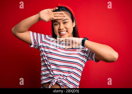 Jeune fille asiatique porte un t-shirt rayé décontracté sur fond rouge isolé sourire gai jouer un joli coup de pied avec les mains montrant le visage. Su Banque D'Images