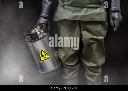 Un homme en costume de protection NBC tient un contenant en métal spécial dans sa main avec un autocollant d'avertissement de danger biologique. Banque D'Images