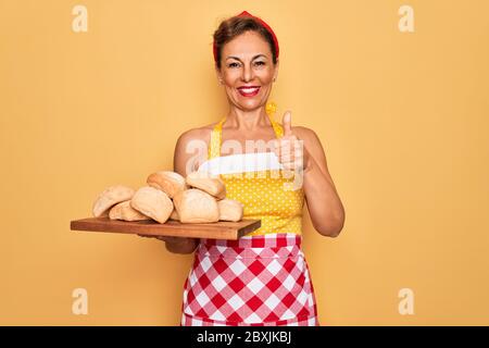 Âge moyen senior épingler femme au foyer porte la robe rétro style 50s et tablier de cuisine pain heureux avec grand sourire faisant signe ok, pouce vers le haut avec la finge Banque D'Images