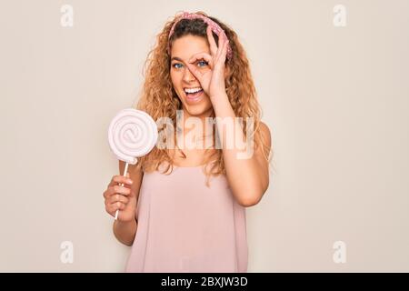 Jeune belle femme blonde avec les yeux bleus manger sucrée bonbon lolipop sur fond rose avec le visage heureux sourire faisant signe ok avec la main sur l'oeil lo Banque D'Images