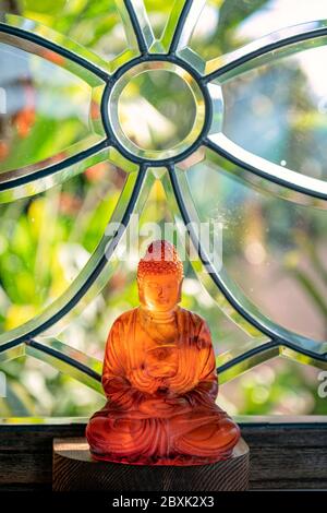 La lumière du soleil brille à travers un Bouddha en verre assis devant une fenêtre avec des plantes en arrière-plan. Banque D'Images