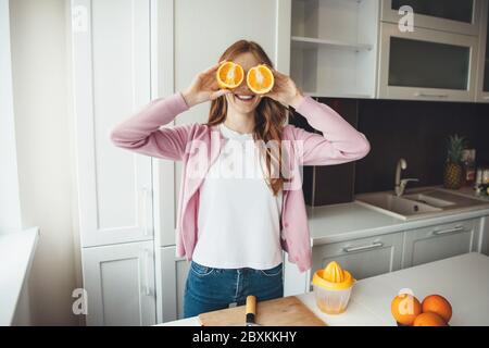 Bonne femme de race blanche avec des cheveux rouges couvrant ses yeux avec des oranges tranchées tout en faisant du jus de fruit Banque D'Images