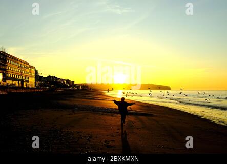 Lever du soleil au-dessus de Culver dans la baie de Sandown bord de mer de Sandown avec une femme courir avec les bras s'écartez vers les pigeons du soleil en vol Banque D'Images