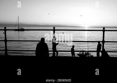 Scène en front de mer au coucher du soleil les gens dans la silhouette des cannes à pêche accroché yacht au-dessus de l'horizon ancré dans la baie calme mer front de mer Banque D'Images