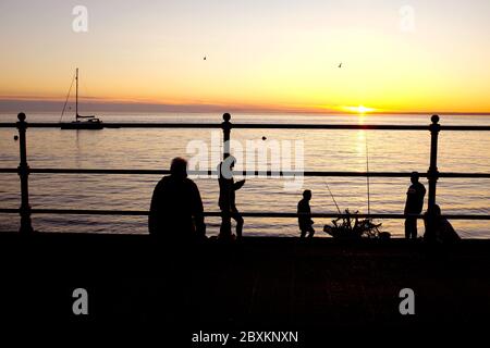 Scène en front de mer au coucher du soleil les gens dans la silhouette des cannes à pêche accroché yacht au-dessus de l'horizon ancré dans la baie calme mer front de mer Banque D'Images