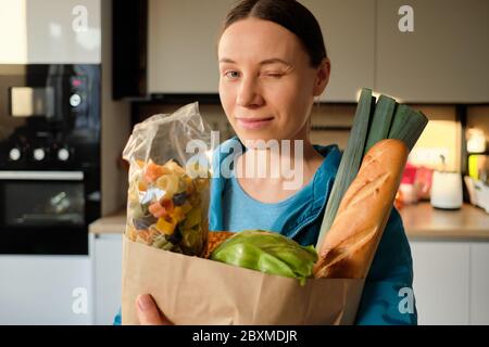 Portrait de la jeune femme dans le sport werar tenant des légumes biologiques, des pâtes et élevés dans un sac de papier artisanal à la maison dans la cuisine Banque D'Images