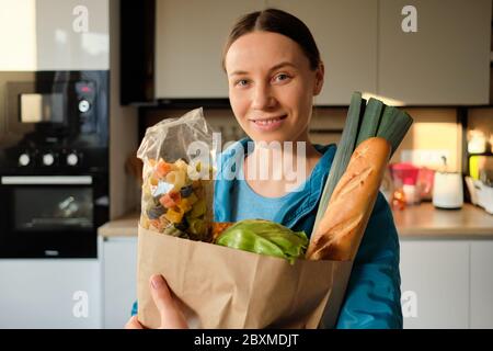 Portrait de la jeune femme dans le sport werar tenant des légumes biologiques, des pâtes et élevés dans un sac de papier artisanal à la maison dans la cuisine Banque D'Images