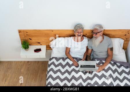 Heureux couple senior regardant des vidéos assis sur le lit tout en riant ensemble - personnes d'âge mûr ayant le plaisir en utilisant PC portable à la maison faisant appel vidéo - Techn Banque D'Images