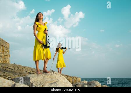 Deux belles sœurs jumelles violinistes dans des robes de concert jaunes jouent des violons électriques sur les rochers au bord de la mer Méditerranée à Antalya, Turquie. Mer Banque D'Images