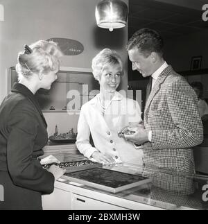 L'amour dans les années 1950. Un jeune couple dans une boutique de bijoux est de choisir des anneaux de mariage. Une employée de vente est debout au comptoir avec une caisse de plusieurs modèles différents d'anneaux. Kristoffersson réf. CG35 Banque D'Images